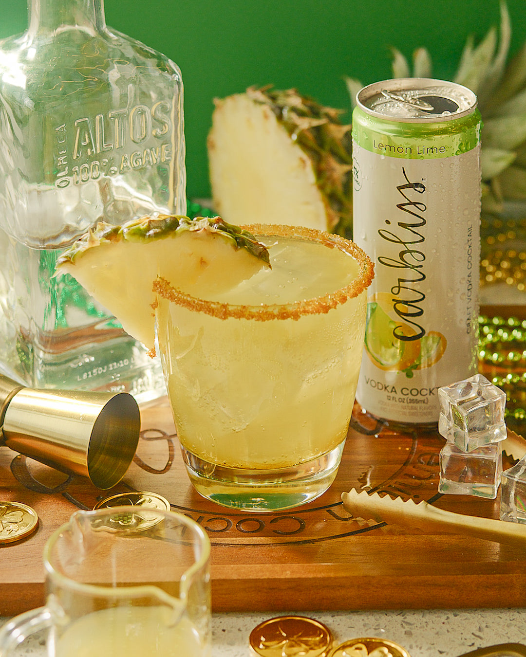 Cocktail with pineapple garnish, bottle, can, and bar tools on a wooden board.